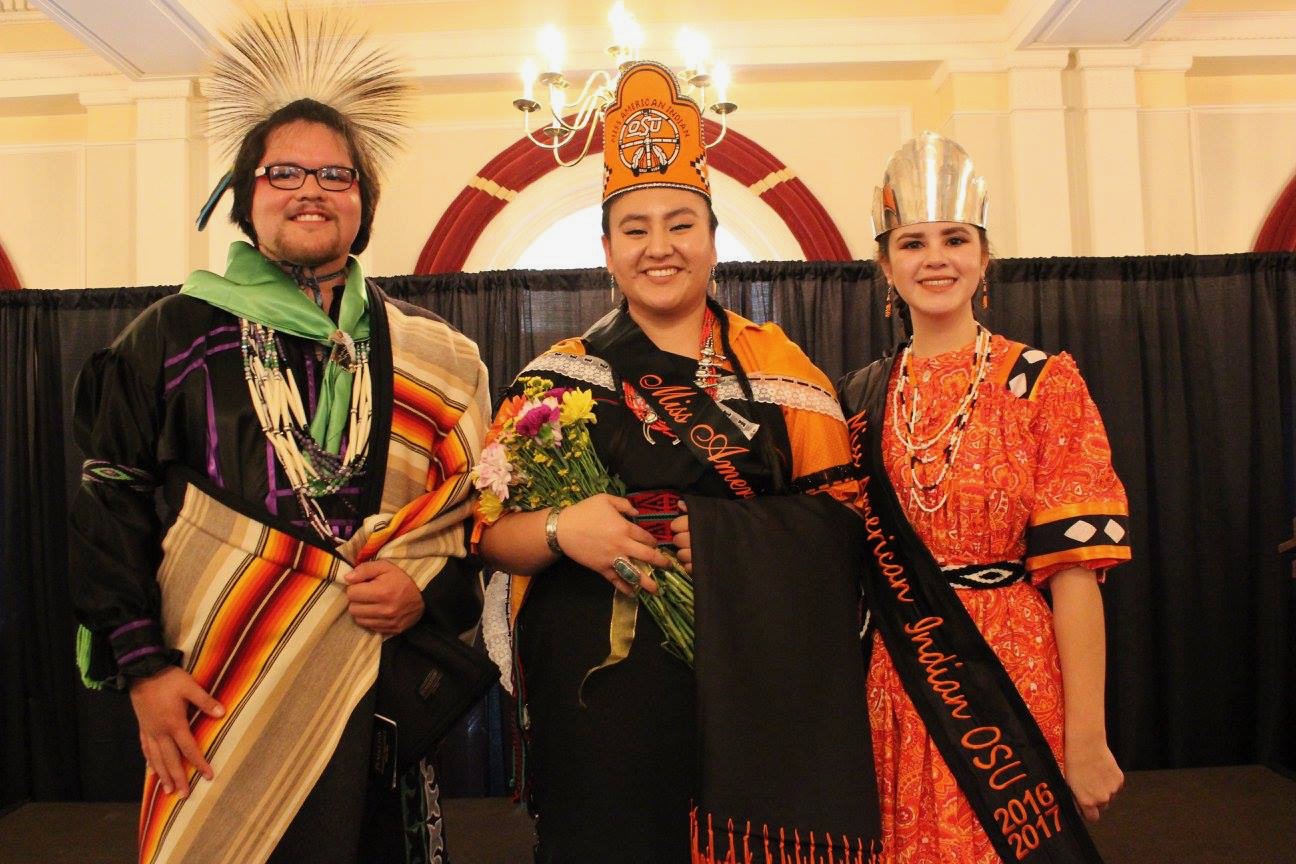 Miss American Indian OSU Cydney, Mister American Indian OSU Che, and a former Miss American Indian OSU pictured together, smiling at the camera. All are wearing sashes and crowns with their traditional clothing Meg.