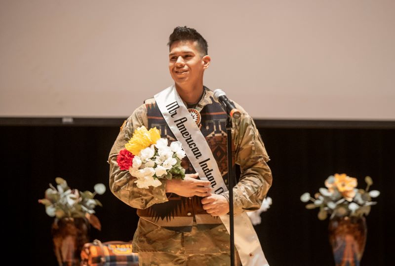 Mister American Indian OSU Luke posing at the MAIOSU pageant, holding flowers and wearing the MAIOSU sash over his Army uniform.