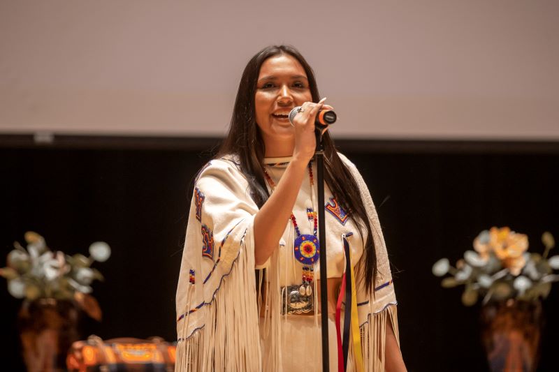 Miss American Indian OSU Gabby pictured speaking into a microphone at the MAIOSU pageant, wearing a traditional buckskin dress.