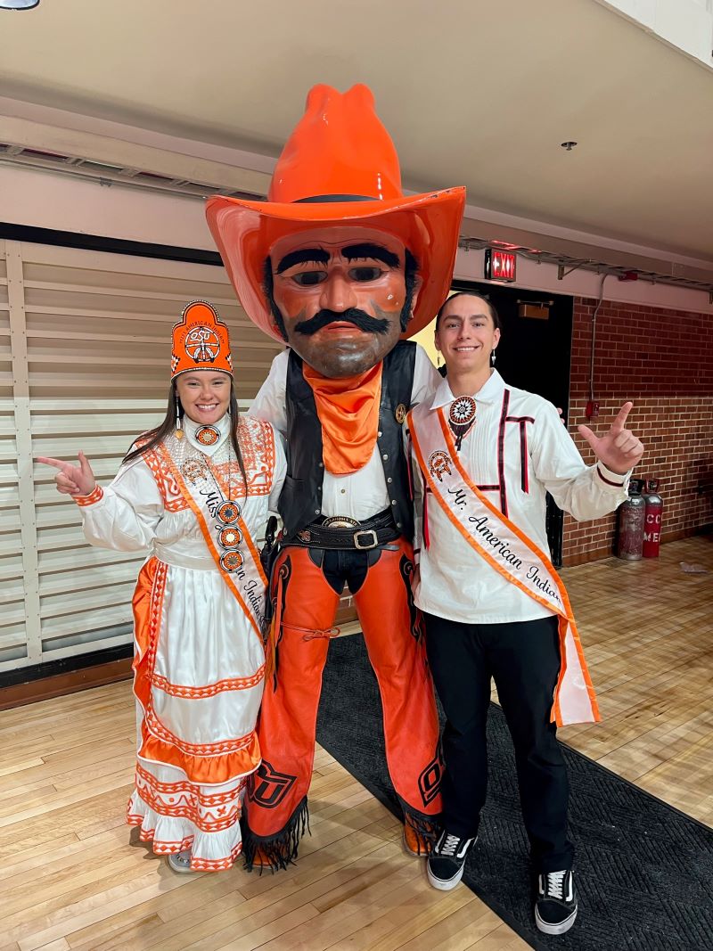 Miss American Indian OSU Shaeleigh and Mister American Indian OSU Caleb, wearing traditional clothing, posing with Pistol Pete at the N7 event.