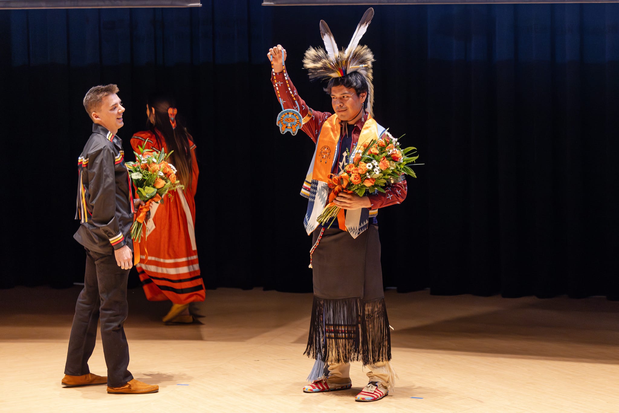 Jaden Waters after winning Mister American Indian OSU 2025, holding up his beaded MAIOSU medallion, wearing a MAIOSU stole and his Kiowa traditional regalia, and holding a bouquet of orange roses.