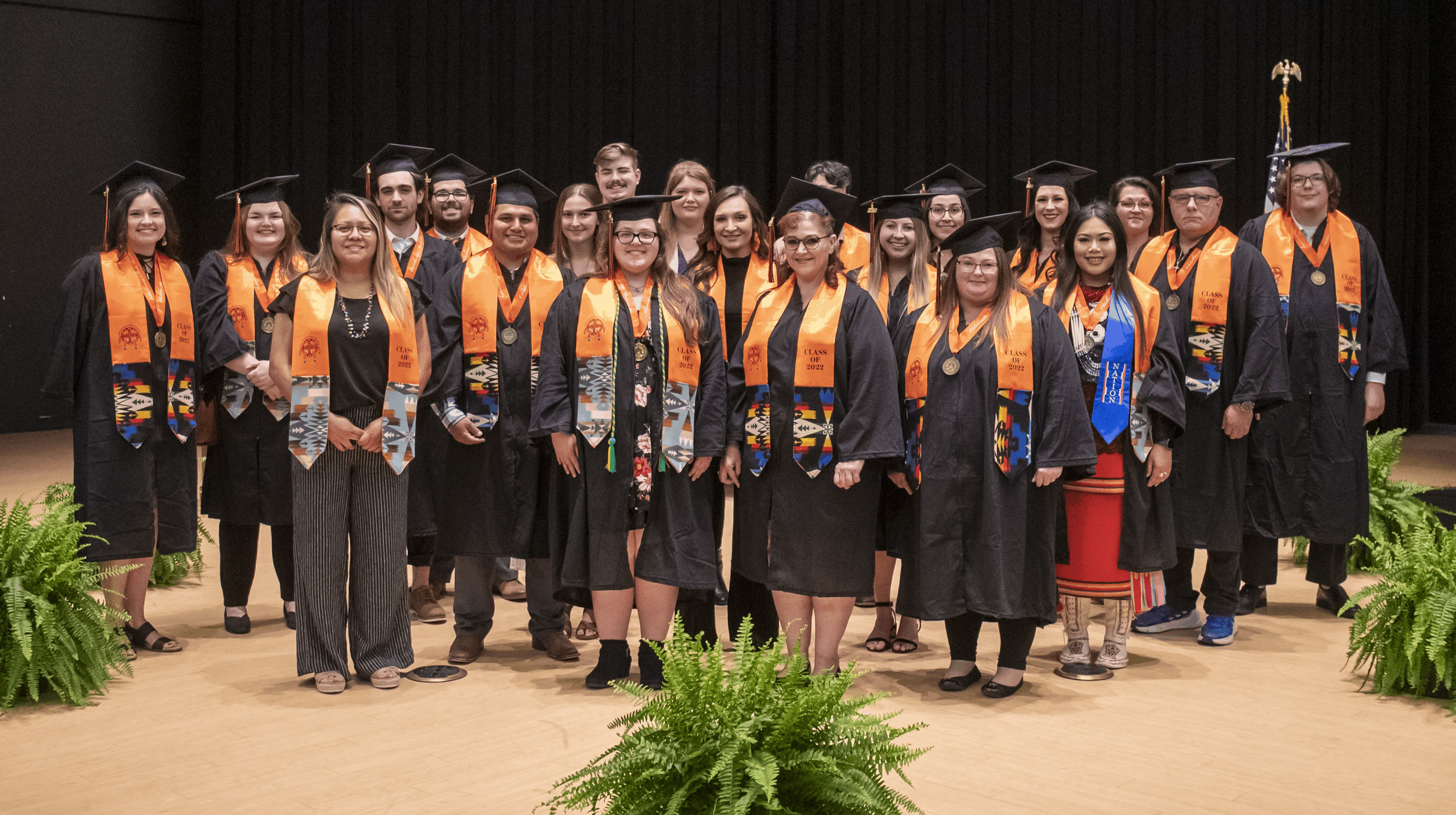 Group of graduates in caps and gowns with orange stoles, standing on a stage.