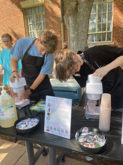 Two people, both wearing black aprons, making snow cones at an outdoor table with supplies and an involvement sign for Stickball and Snowcones for the Center's 9th Birthday.jpg